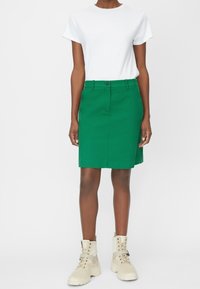 Jupe en coton vert à coupe droite, avec poches avant et passants de ceinture, associée à un t-shirt blanc et des bottes beiges à lacets.