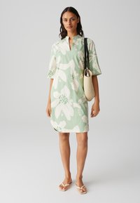 Light green floral dress with short sleeves, a stand collar, and large white petals with green accents. Paired with a beige tote bag.