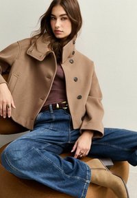 Manteau cropped en mélange de laine marron avec col montant et grands boutons, associé à un jean bleu à jambes larges et des bottines beiges. Texture lisse.