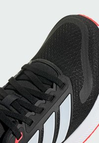 Chaussure de sport noire avec un dessus en maille, des lacets ronds, des accents blancs et une semelle texturée avec un détail rouge au talon.