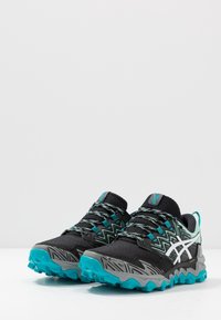 Chaussures de trail de course noires et teal avec une tige en mesh texturé, un motif en zigzag et une semelle extérieure à traction robuste. Comprend des lacets et des accents du logo.