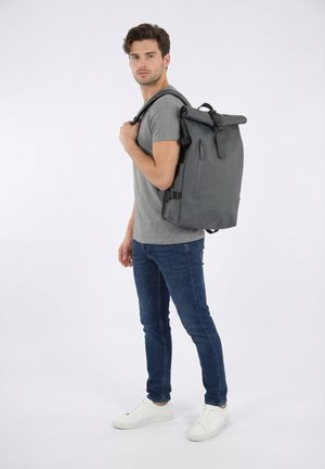 Giovane uomo con t-shirt grigia e jeans che indossa e tiene uno zaino grande grigio con chiusura a rotolo sulla schiena, in piedi su sfondo bianco uniforme.