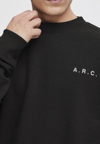 Sort sweatshirt lavet af bomuld, med rund halsudskæring, ribbede manchetter og trykt tekst "A.R.C." i hvidt på venstre brystområde.