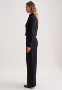 Zwarte blouse met lange mouwen en een staande kraag, gecombineerd met zwarte rechte broek. Glad stof en op maat gemaakt ontwerp met knoopdetails.