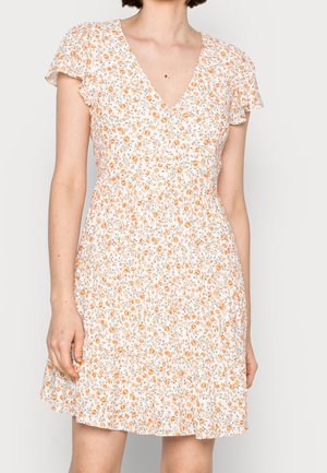 Kobieta ubrana w białą sukienkę z krótkim rękawem z pomarańczowym kwiatowym wzorem, dekoltem w serek i falbankowymi rękawkami, stojąca na jednolitym tle.