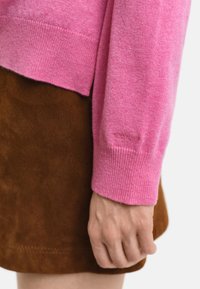 Rosa gestrickter Pullover mit gerippten Bündchen, über einem braunen Wildlederrock getragen. Auffälliger Texturunterschied zwischen den Materialien. Markenlogo am Bündchen.