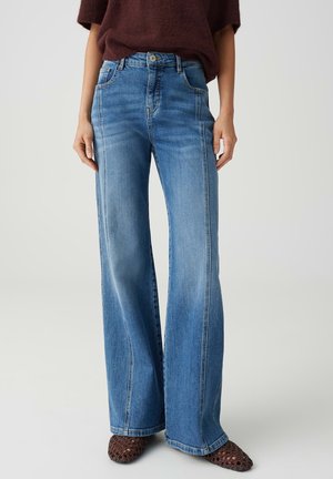 Femme portant un jean bleu taille haute à jambes larges avec des détails de coutures, des chaussures tressées marron et un haut marron à manches courtes, debout devant un fond uni.