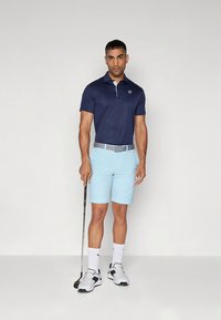 Golfpolo i marineblå med hvit krage, kombinert med lyseblå shorts, grått belte og svarte og hvite golfsko, holder en putter.