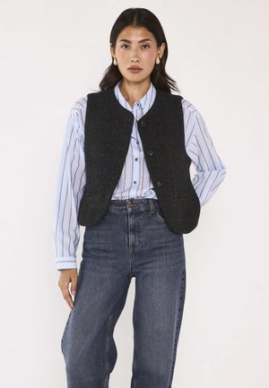 Woman wearing black buttoned vest over light blue striped shirt and high-waisted dark blue jeans, standing against plain background.
