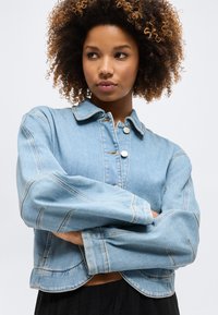 Νεαρή γυναίκα με σγουρά μαλλιά φορά ένα ανοιχτό γαλάζιο τζιν μπουφάν με μεταλλικά κουμπιά, με σταυρωμένα τα χέρια, κοιτάζει στο πλάι μπροστά σε μια απλή φόντο.