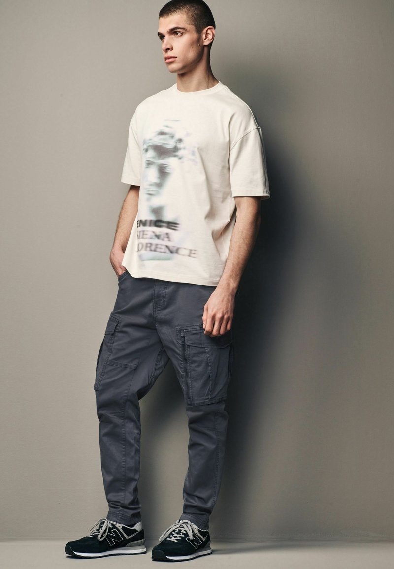 Creme oversized t-shirt med et sløret grafisk tryk, parret med slim-fit grå cargo bukser med sidelommer og sneakers.