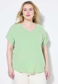 Maglietta verde chiaro a V con maniche corte, realizzata in tessuto testurizzato con un motivo a griglia. Vestibilità ampia con orlo arrotondato.
