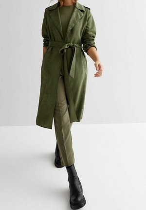 Persoon loopt, gekleed in een olijfgroene trenchcoat met riem, bijpassende broek en zwarte chunky enkellaarzen tegen een effen lichte achtergrond.