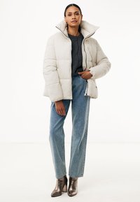 Giubbotto imbottito beige chiaro con collo alto, design trapuntato e chiusura frontale a zip. Abbinato a jeans in denim blu e stivali con tacco metallici.