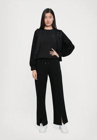 Zwarte casual outfit bestaande uit een lange mouwen pullover en wijde broek met zijsplitten, aangevuld met witte sneakers. Glad stoftextuur.