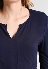 Top azul marino con escote en V, mangas largas y dos bolsillos frontales. Hecho de una tela suave y ligera con un diseño simple y limpio.