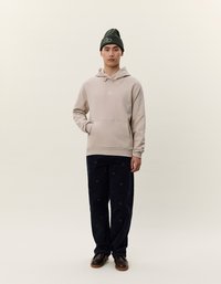 Beige hettegenser med frontlomme, kombinert med mørk marineblå bukser med sirkulære mønstre, båret med en mørkegrønn lue og brune sko.