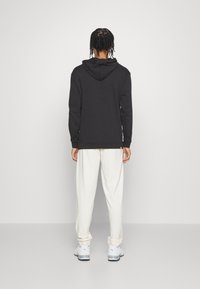 Person med flätat hår iförd en svart hoodie, vita lösa byxor och vita sneakers, står vänd mot en enkel vit bakgrund.