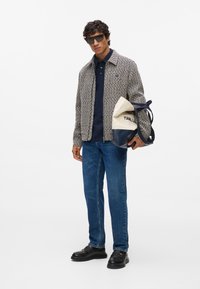 Chaqueta de tejido en crema y azul marino, camiseta polo azul, vaqueros azules y zapatos negros. Sosteniendo un bolso en crema y azul marino con un logo impreso.