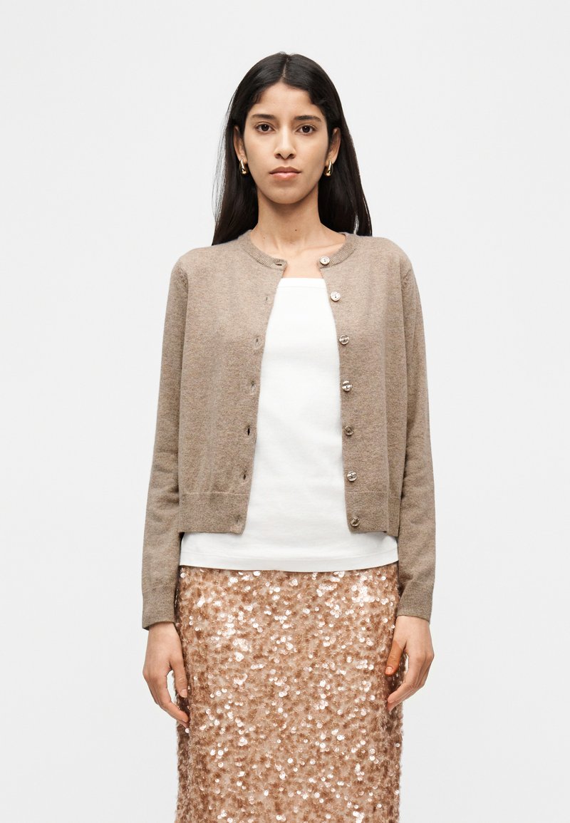 Καφέ πλεκτή ζακέτα με στρογγυλή λαιμόκοψη, μπροστινά κουμπιά και μακριά μανίκια, συνδυασμένη με λευκό τοπ και φούστα σε ροζ χρυσό με παγιέτες.