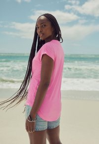 Ružičasta majica kratkih rukava s okruglim izrezom u kombinaciji s traper kratkim hlačama. Osoba ima dugu pletenicu i okrenuta je leđima prema kameri.