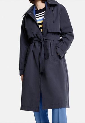 Femme portant un trench-coat bleu marine ceinturé par-dessus un pull rayé et un pantalon bleu clair, debout devant un fond blanc uni.