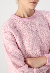 Osoba w jasnoróżowym swetrze z teksturowanym splotem, z krzyżującymi się ramionami, pokazana od szyi do talii na jednolitym tle.