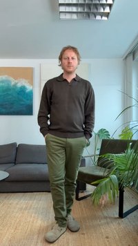 Mężczyzna stojący z rękami w kieszeniach w salonie z szarą sofą, zielonym krzesłem, roślinami doniczkowymi i abstrakcyjną sztuką na ścianie.