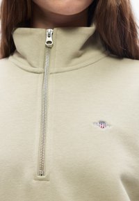 Μπεζ ζακέτα με φερμουάρ και ψηλό γιακά που φοριέται από άτομο, με μικρό κεντητό λογότυπο με ασπίδα και αστέρια στο στήθος.