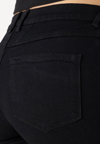 Jeans de mezclilla negra con una vista en primer plano del bolsillo trasero. Presenta un corte recto y una textura sutil. Detalles de costura visibles en el bolsillo.