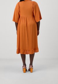 Femme debout portant une robe orange mi-longue avec des manches bouffantes et des sandales à talons blocs corail sur un fond uni.