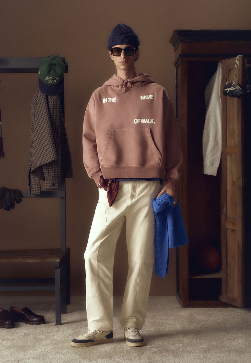 Hooded dukserica u zamućenoj ružičastoj boji s bijelim tekstom, kreme široke hlače, bež i sivi tenisice, te plavi pleteni dodatak.