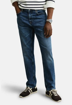 Mann trägt blaue Jeans, schwarz-weiße Turnschuhe, gestreiftes Langarmshirt und Armbanduhr, mit einer Hand in der Tasche.