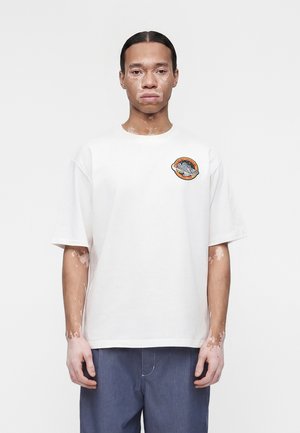 Osoba s vitiligom stojaca, oblečená v bielom tričku s oranžovo-čiernym kruhovým nášivom na ľavej strane hrudníka a modrých nohaviciach.