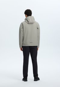 Veste imperméable grise avec une capuche ajustée, texture lisse, manches longues et accents réfléchissants ; portée avec un pantalon et des chaussures noires.