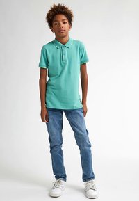 Turkos pikétröja med krage, två knappar och gröna detaljer; kombinerad med blå jeans och vita sneakers; modellen står upprätt.