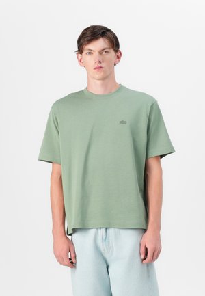 T-shirt vert à manches courtes en tissu doux avec un col rond. Il présente un logo minimaliste sur la poitrine gauche et une coupe décontractée.