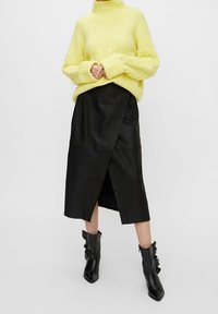 Pull à col roulé jaune en tricot à texture épaisse, assorti à une jupe portefeuille en cuir noir avec une fente latérale et des bottines noires.
