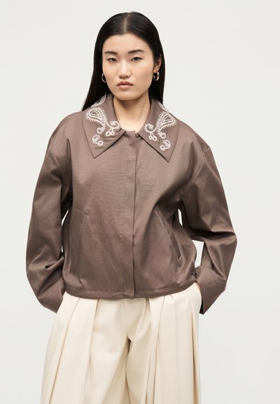 Γυναίκα με ίσια μαύρα μαλλιά που φοράει καφέ σακάκι με λευκή κεντητή γιακά και εκρού πλισέ παντελόνι με φαρδιές μπατζάκια.