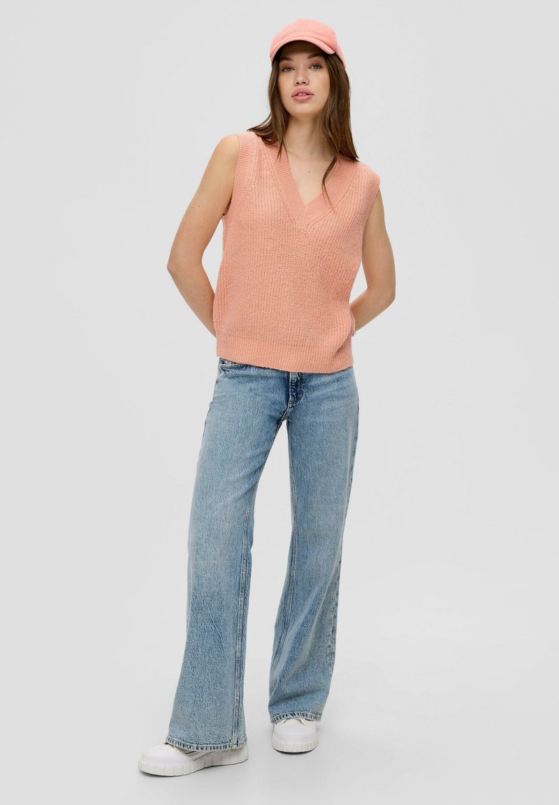 Pfirsichfarbener, ärmelloser Strickpullover mit V-Ausschnitt, kombiniert mit hellblauen, weit geschnittenen Jeans und weißen Plateausneakern. Eine Mütze rundet das Outfit ab.