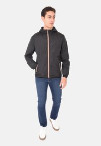 Giacca leggera nera con cappuccio, con dettagli della cerniera arancioni. Abbinata a jeans blu e scarpe da ginnastica bianche. Texture liscia.