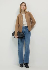 Donna in giubbotto di suede beige, top bianco, jeans a vita alta blu, stivaletti neri alla caviglia, che tiene una grande pochette nera contro uno sfondo neutro.
