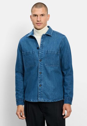Mann, der ein blaues Jeanshemd mit Knöpfen über einem weißen Rollkragenpullover trägt, steht vor einem einfarbigen hellen Hintergrund.