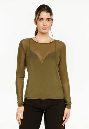 Femme aux cheveux foncés attachés, portant un haut transparent à manches longues vert olive par-dessus un bralette assorti et un pantalon noir, debout devant un fond blanc.