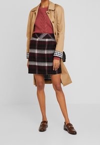 Beige trenkkjole over en burgunder kabelstrikket genser og rutete skjørt i rødt, svart og hvitt. Brune loafers med hardware-detalj.