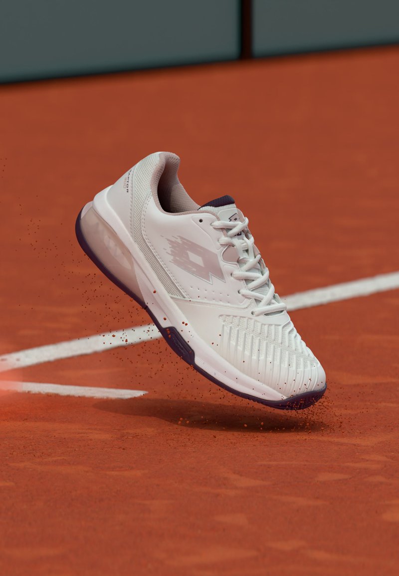 White tennis shoe in mid-air kicking up red clay on a tennis court near a white boundary line.