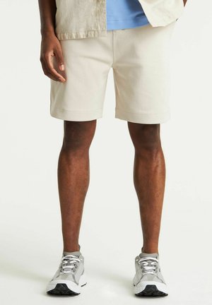 Shorts en coton beige avec une coupe classique et des coutures visibles. Portés avec des chaussures de sport grises présentant des accents blancs et noirs.
