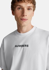 Mladý muž s krátkými kudrnatými vlasy, oblečený v bílém tričku s tučně vytištěným slovem "ČÍSLA" na hrudi.