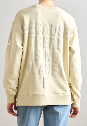 Personne portant un sweat crème avec un grand texte en relief au dos indiquant « "Because there is no Planet B," », associé à un jean bleu.