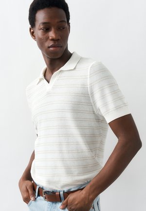 Joven con las manos en los bolsillos, vistiendo una camiseta polo de manga corta blanca con rayas y vaqueros azul claro sobre un fondo liso.